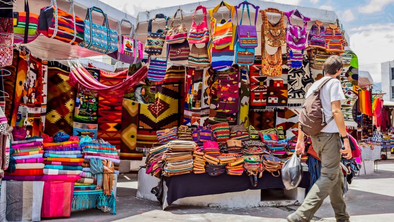 Young traveler with backpack walks past brightly colored market stall with woven goods on the Otavalo Escape tour in Ecuador.