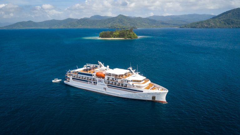 Aerial view of a small white expedition ship cruising a Papua New Guinea cruise in deep blue water beside green islands.