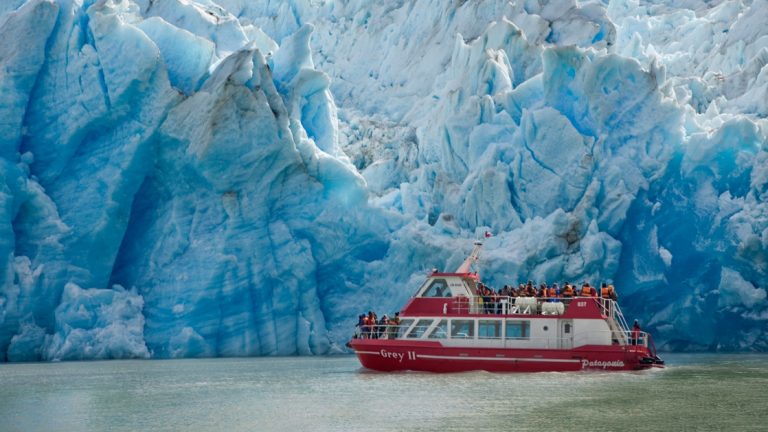 Red & white boat cruises beside a massive blue glacier in Torres del Paine National Park, Patagonia, Chile.