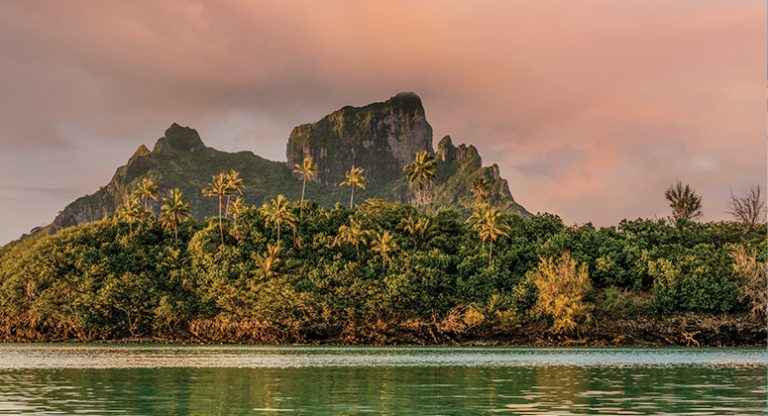 Tropical island with rocky peaks & palm trees, seen during a pink sunset on the Pearls of The Pacific Society & Tuamotu Cruise