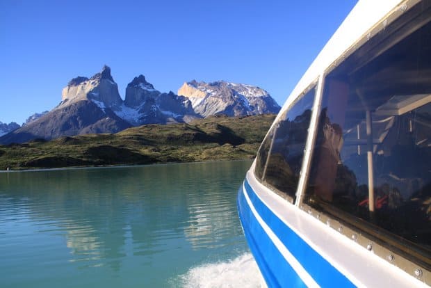 Ferry crossing on a lake moving towards a rocky shoreline with large mountains behind and clear blue sky.