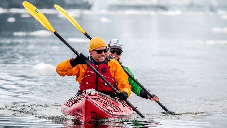 Tandem kayakers paddle in sync in a red kayak with yellow paddles & bright-colored clothes in calm water in Antarctica.