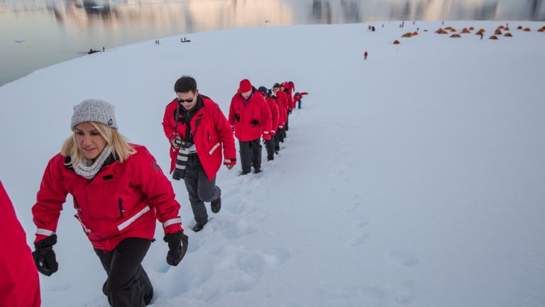 Group of hikers in red jackets walks up a snowfield away from tents at dusk on the Quest For The Antarctic Circle cruise.