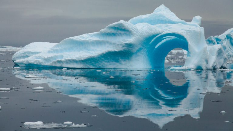 Large blue iceberg with a tunnel rises out of glassy water on a cloudy day on the Quest For The Antarctic Circle cruise.
