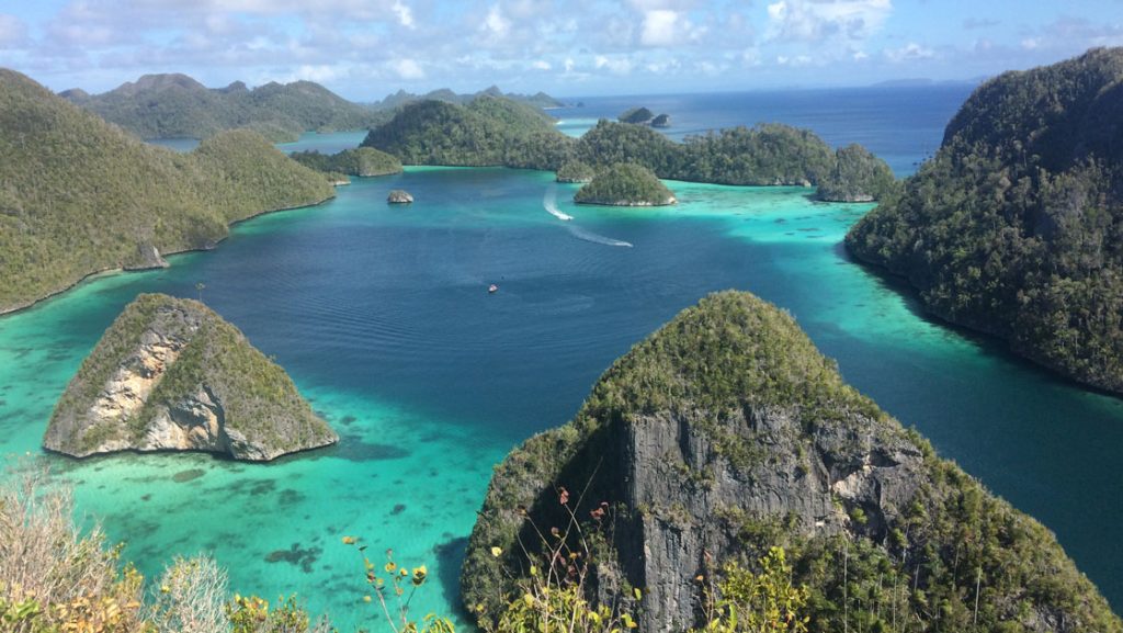 looking down at an indonesia archipelago scattered around turquoise water with a small ship traveling in the distance on the Spice Islands and Raja Ampat cruise
