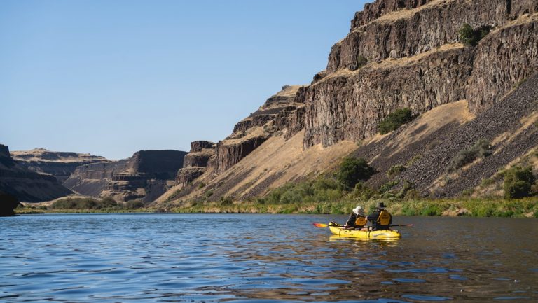 Guests in a yellow double kayak paddle down a river gorge, surrounded by steep, rocky walls.