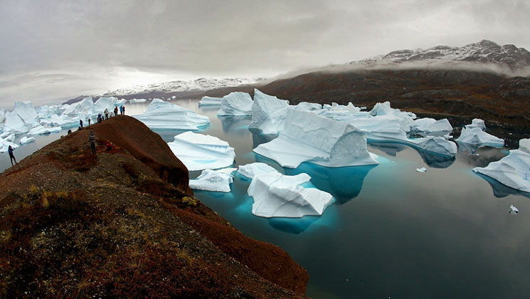 Hikers overlook large icebergs sitting in Arctic waters on a cloudy day during the Scoresby Sund Aurora Borealis voyage.