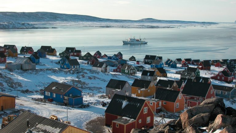 Aerial view of small seaside town in the Arctic with colorful homes & small ship sitting offshore, on a sunny day.