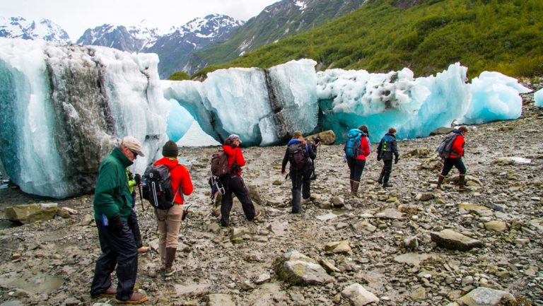 As part of a shore excursion, a group of travelers walks past large teal beached icebergs that washed ashore in Alaska.