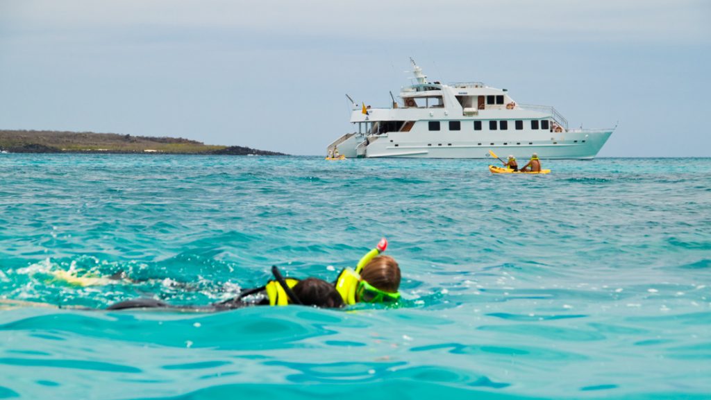 2 Galapagos travelers snorkel in turquoise water as 2 more paddle a yellow kayak in front of Seaman Journey white catamaran.