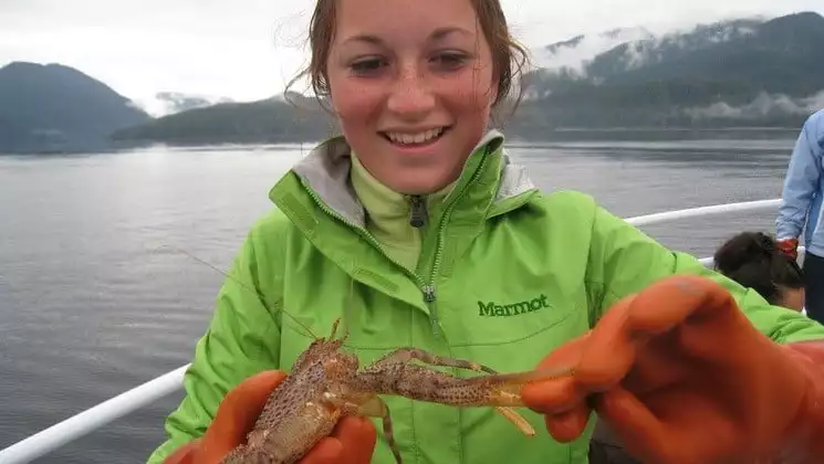 girl in a green jacket holds a crayfish in alaska with calm water and forest behind her