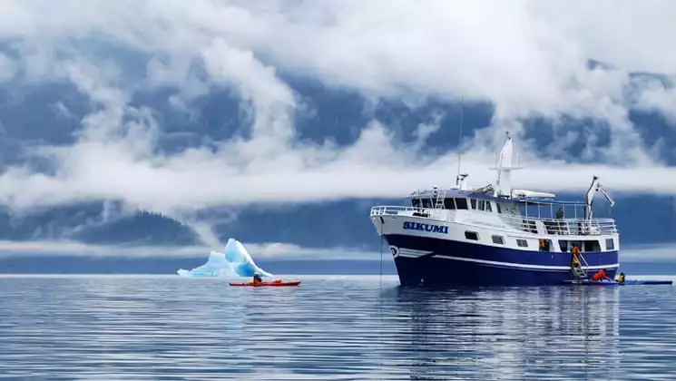 sikumi small ship anchored while an adventure traveler kayaks in front of an iceberg on a misty day in alaska
