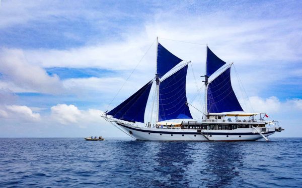 A wooden Indonesia small cruise ship is shown on starboard side with blue sails up with blue ocean, blue sky and white clouds