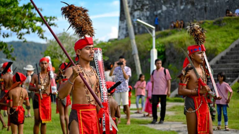 Men in red loincloths and red headbands with tall brown feathers march in order beside a stone fort on a sunny day during the Spice Islands & Raja Ampat small ship cruise in Indonesia.