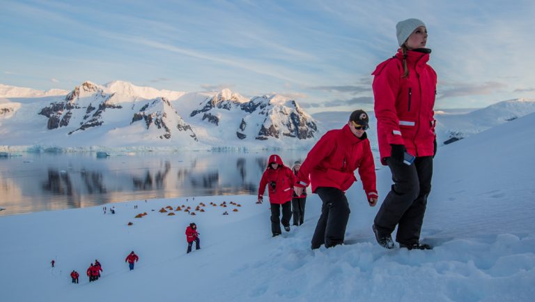 Group of hikers in red jackets walks up snowy hill, away from colorful tents, at dusk on Spirit of Shackleton Aboard Expedition cruise.