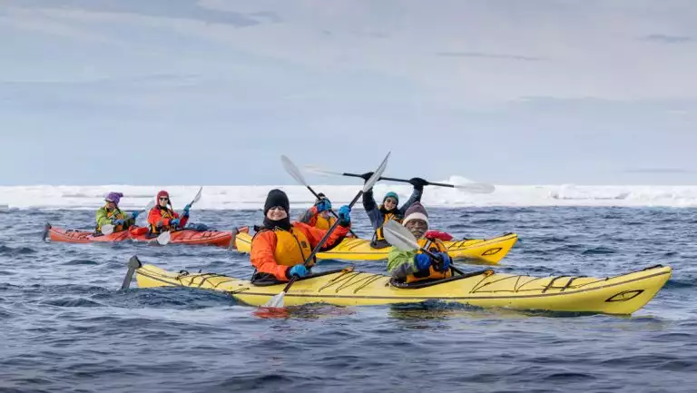 A few tandem kayakers dressed warmly paddle yellow & red boats in calm sea beside floating ice during a Svalbard cruise.