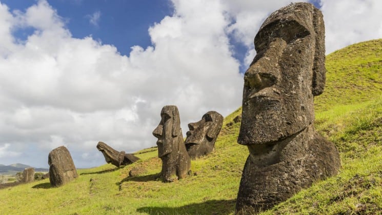 a grassy hillside in easter island chile with several moai statues sitting on it with puffy clouds behind them
