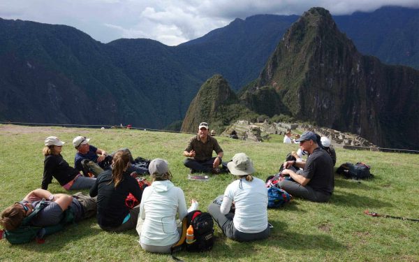 Group of travlers sitting on the grass at Machu Picchu listening to a guide.