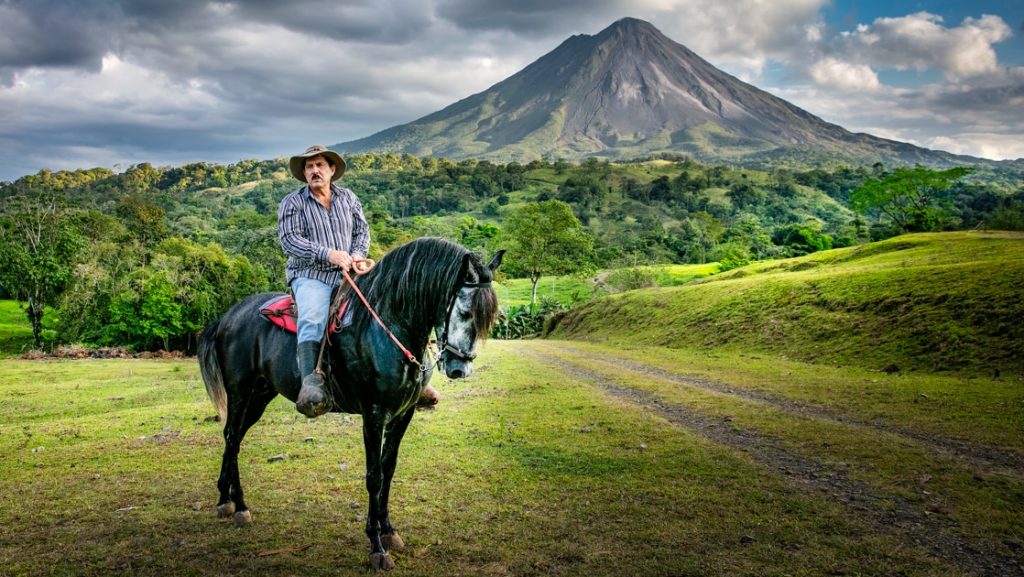Man on black horse stands in a green field with bright green forest & Costa Rica's Arenal volcano in the background.
