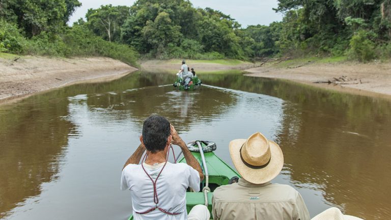 Two male travelers in a skiff follow another skiff on the Amazon River. Seen from their backs one man holds binoculars and the other wears a hat.