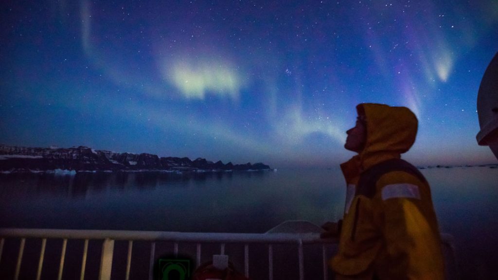 Arctic traveler in yellow polar jacket stands out on deck of ship watching northern lights of purple, blue & green in Greenland.