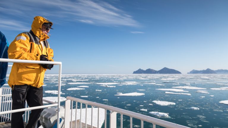 Arctic traveler in yellow polar parka stands on deck of ship looking at sea with floating ice chunks on a northern lights cruise.