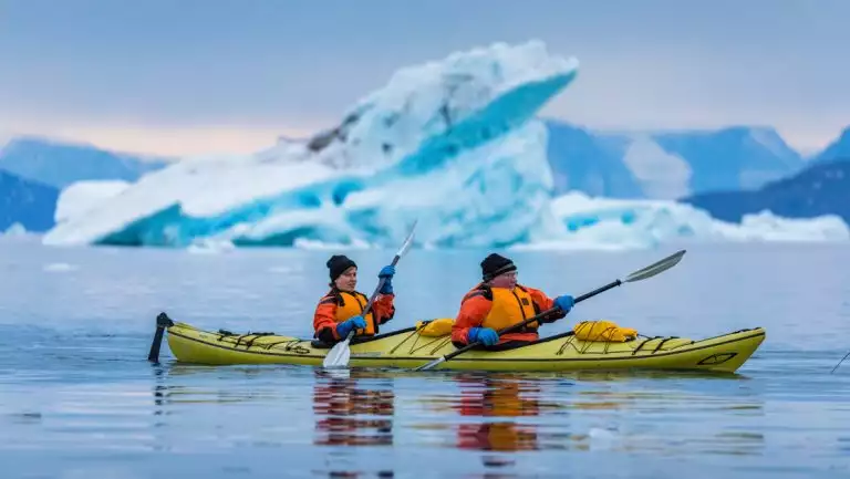 Tandem kayakers in lime green boat paddle glassy water beside a large blue iceberg during an Under The Northern Lights cruise.
