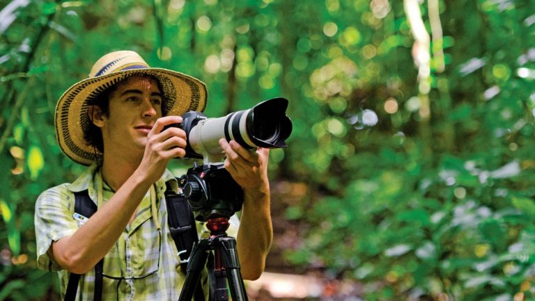 Male traveler uses a large camera on a tripod in the lush green Belize jungle, during the Wild Belize Escape small ship cruise.