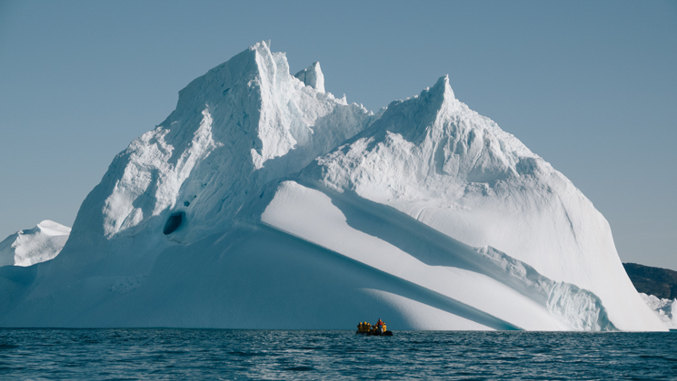 Zodiac boat with south Greenland travelers cruises past a massive iceberg resembling a snow-covered mountain in the sun.