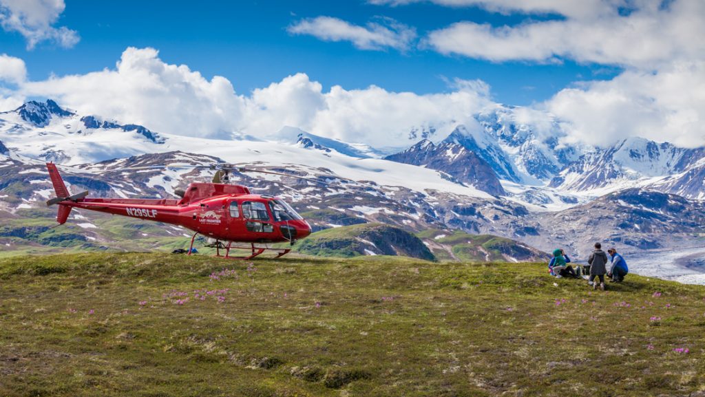 Red helicopter sits turned off atop a high meadow with snowy peaks beyond as Within the Wild Alaska guests have a picnic lunch.