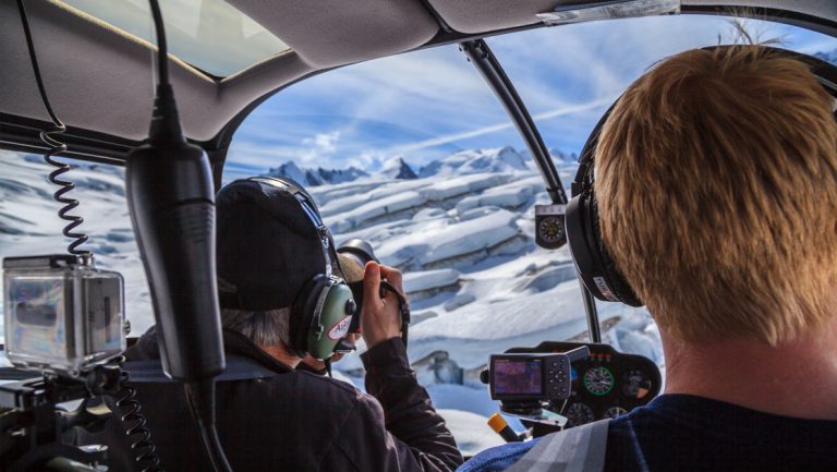 Man photographs glacier ice from the front of a helicopter while pilot flies on a sunny day during the Within the Wild Alaska tour.