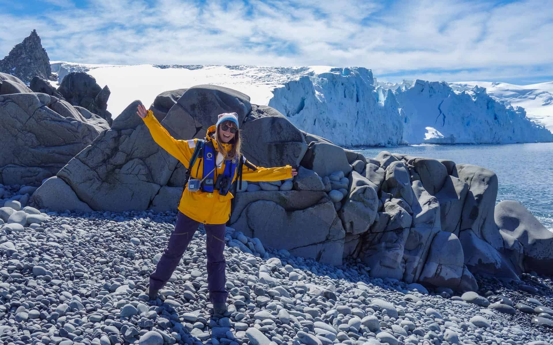 Young woman excitedly standing on a rocky beach in Antarctica with bright yellow jacket and massive icebergs behind her.