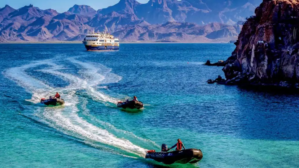 3 black inflatable Zodiac boats cruise away from Nat Geo Venture ship through turquoise waters near rocky coastline in Baja.
