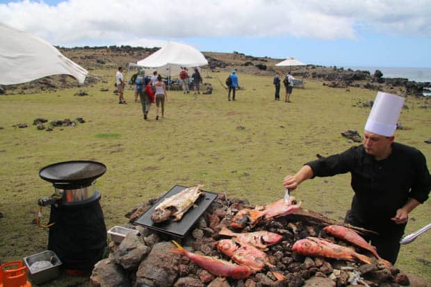 A chef preparing whole fish in a field with people and tents.