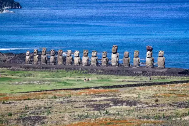 A line of Moai statues with the ocean in the background with visitors in the grass.