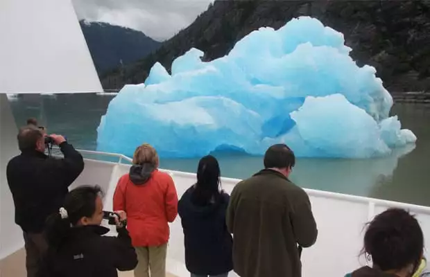 group of alaska small ship cruise travelers on the side of the boat taking pictures of a large iridescent blue piece of floating ice