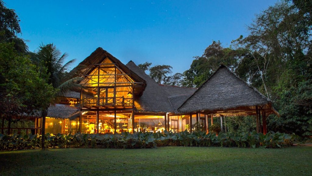 The exterior of the Inkaterra Reserva Amazonica at night, with thatched roofs and lights. It is an eco-luxury lodge in the heart of the Peruvian Amazon Rainforest, adjacent to the Tambopata National Park