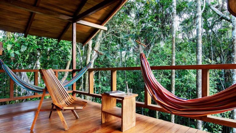Private balcony at Sacha Jungle Lodge, with wooden floors & railing, 2 brightly-colored hammocks, wooden chair & book on a small table.