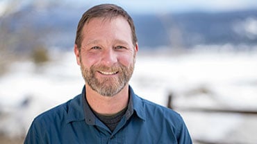 adventuresmith explorations director of sales and operations justin massoni wearing a blue shirt smiling on a sunny day with snow and mountains in the background