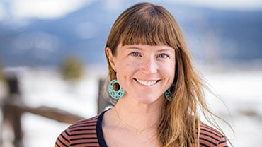 adventuresmith explorations director of content and marketing lis korb smiles wearing a striped shirt on a sunny day with snow and mountains behind her