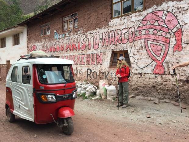 Small red and white 3 wheeled tuk tuk car riding on a dirt street with a traveler standing next to a building with red grafitti.