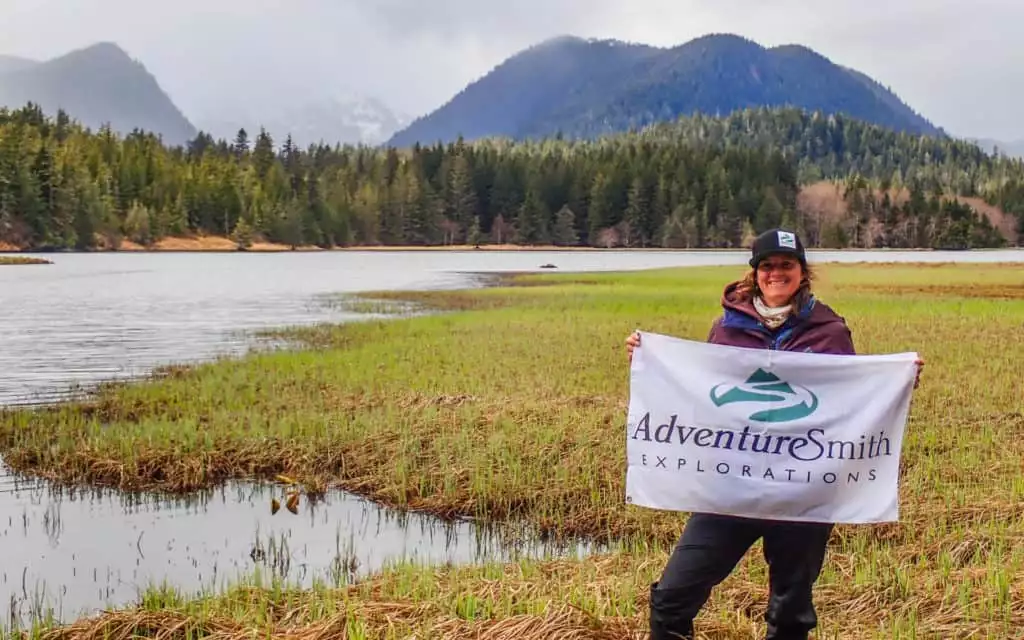 An AdventureSmith traveler with flag in front of a lake and mountains in Alaska.
