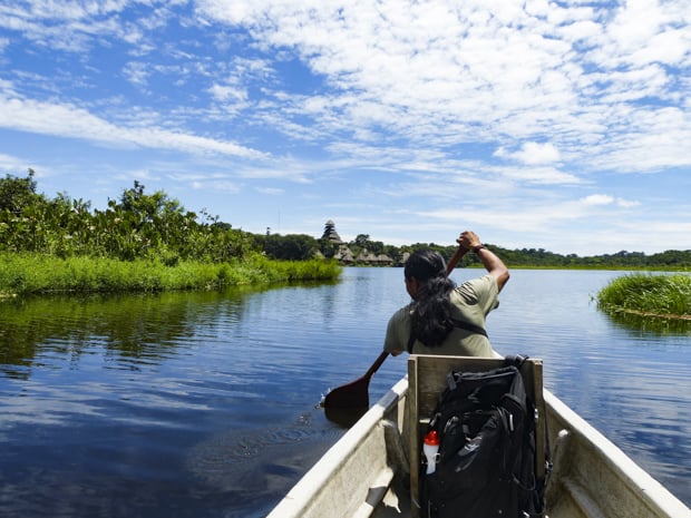 Amazon traveler canoeing on the river towards thatched hut lodge with green grassy water line.