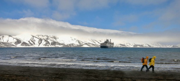 Two passengers on a land excursion in Antarctica walking along the shore with their small ship in the background. 