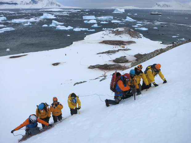 People on a mountaineering excursion in Antarctica climbing uphill on snow with ice picks and ropes with their small ship in the background. 