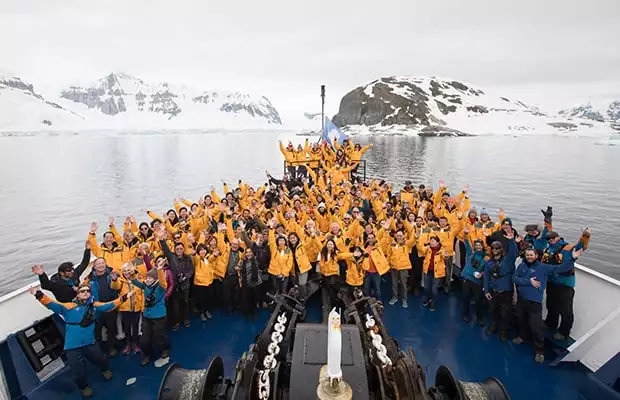 Entire crew and guests posing with their hands up in the air on the bow of their small ship in Antarctica. 