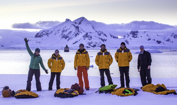 Antarctica camping excursion guests pose next to their bivy sacks on the snow with their small ship cruise leaving in the distance.