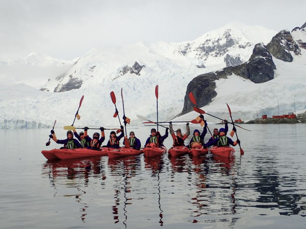 A group Antarctica travelers kayaking in red kayaks with their paddles up and snow and a glacier behind them.