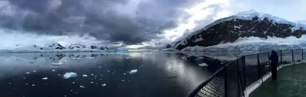 A panorama of the bow of the ship coming into Antarctica with sea ice and mountains and lots of dark clouds.