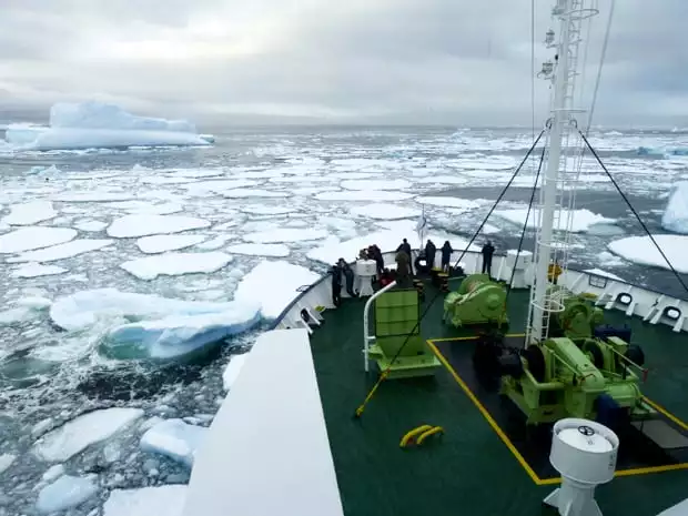 People on the bow of their small ship watching as the ship breaks through some ice in Antarctica. 